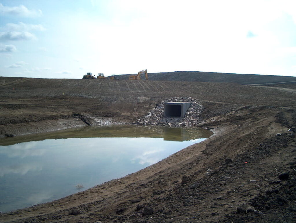 A drainage culvert surrounded by rocks leading into a water retention pond on a construction site with heavy machinery in the background.