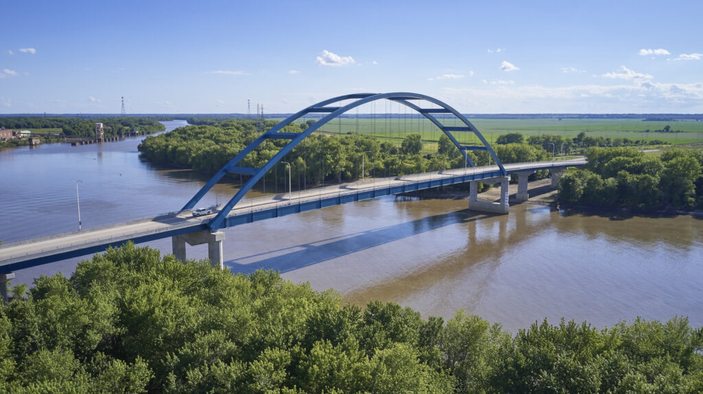 An arched bridge spans over a wide river, with lush green trees on both sides and clear blue skies overhead.