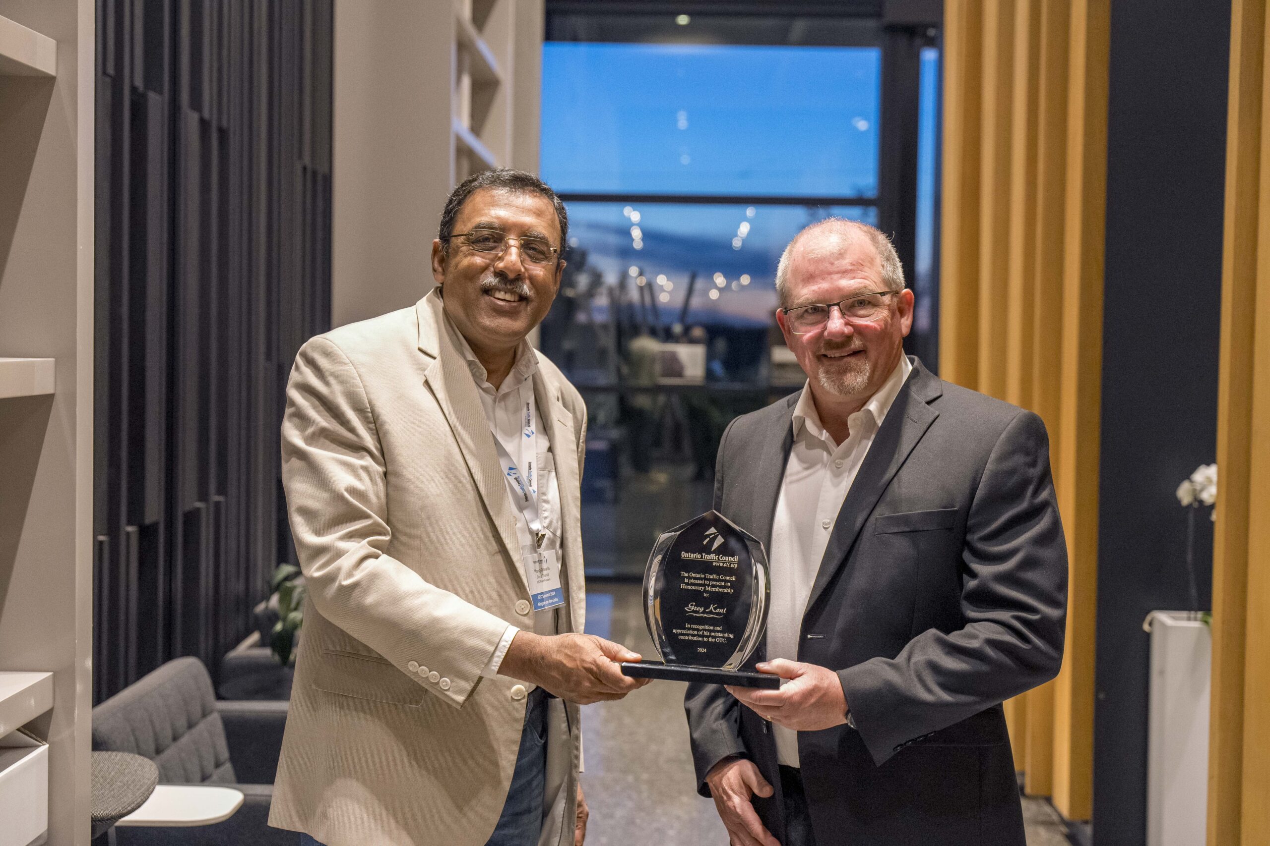 Two men, both wearing suits, are standing indoors. One is holding an award plaque, and they are smiling at the camera. In the background, there are shelves and a window with a view outside.