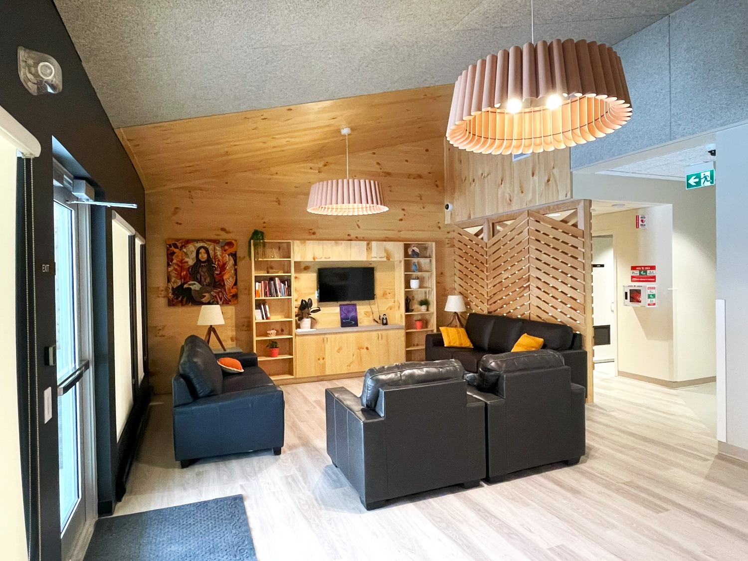 A cozy lounge with black leather sofas, wooden bookshelves, and a wall-mounted TV. Two pendant lights hang from the ceiling. The walls have wooden paneling, and there's a doorway leading to another area.