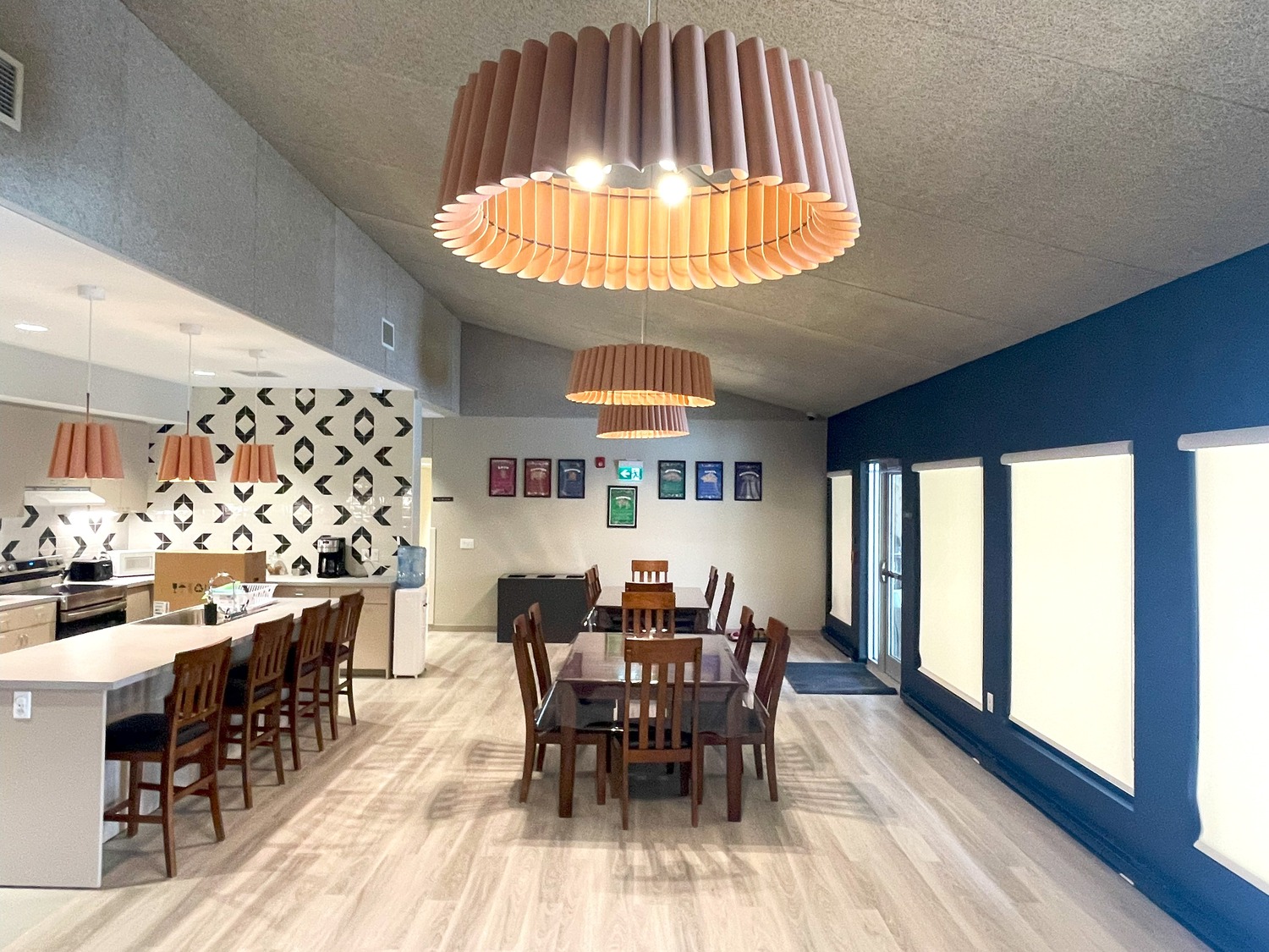 A modern dining room with a long rectangular table and wooden chairs, pendant lights above, and a kitchen area in the background featuring a patterned backsplash and wooden barstools.