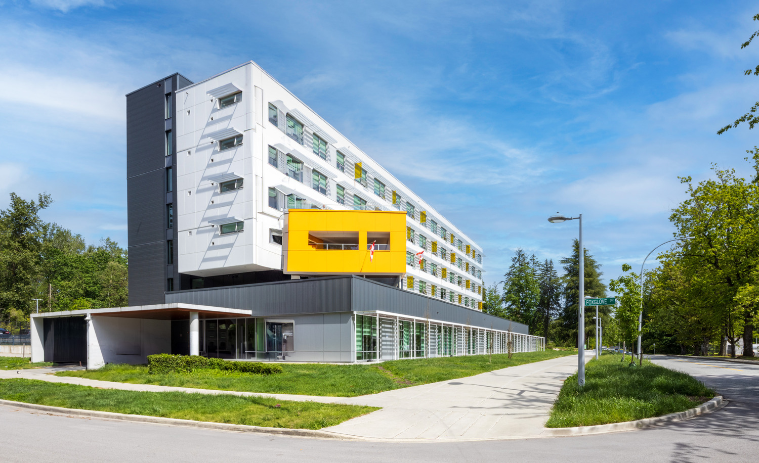 Modern multi-story building with white and gray exterior, featuring a prominent yellow section, large windows, and surrounded by greenery under a clear blue sky.