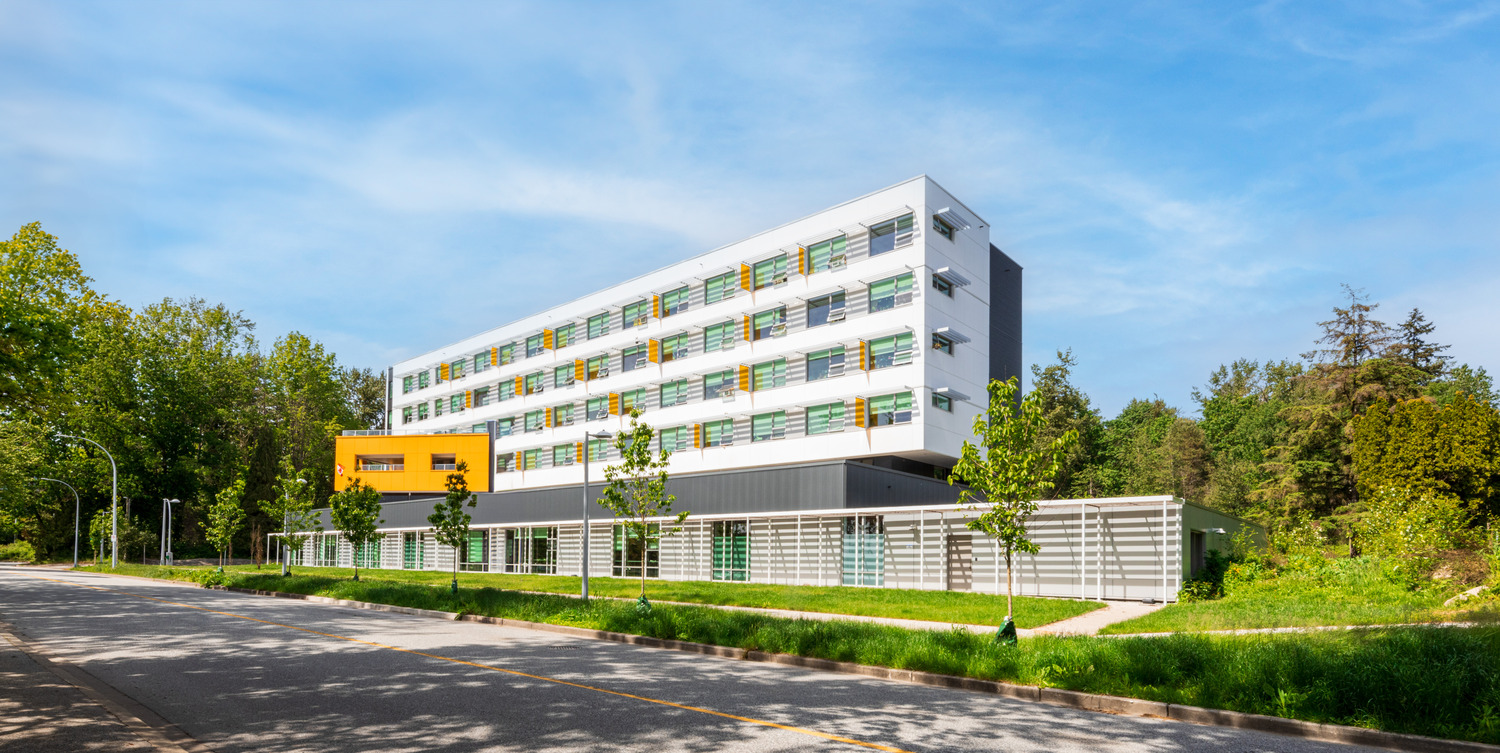 Un bâtiment moderne de plusieurs étages aux accents verts et jaunes se dresse sur un ciel bleu clair. Il est entouré d'arbres et de verdure, avec une route et un trottoir devant.