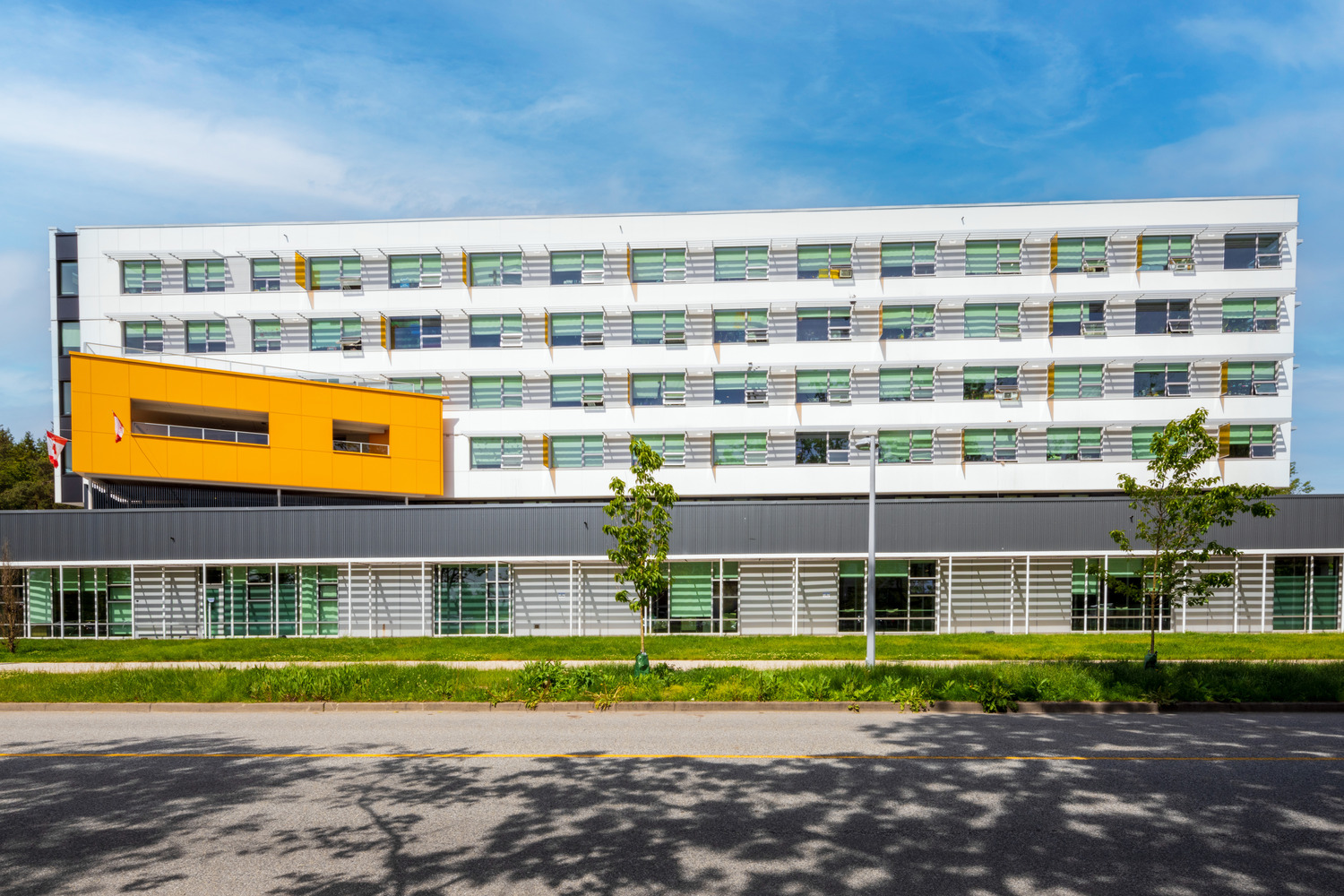 Bâtiment moderne de plusieurs étages avec extérieur blanc et orange, bordé de fenêtres horizontales, sur fond de ciel bleu. De grands arbres et une zone herbeuse sont au premier plan avec une route.