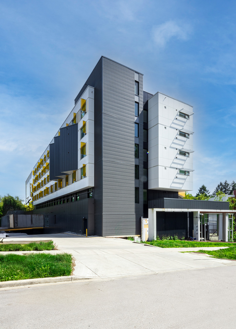 A modern, multi-story building with gray and white exterior, featuring yellow window accents and balconies, stands against a clear, blue sky. A paved driveway and portions of a green lawn are visible.