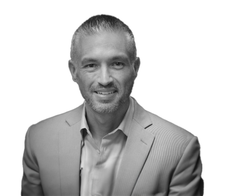 Black and white photo of a man with short, neatly styled hair, wearing a light-colored blazer and shirt, smiling directly at the camera against a white background.