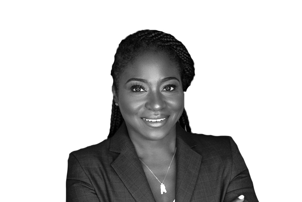 Black and white image of a person wearing a blazer and a necklace, smiling at the camera, with their arms crossed.