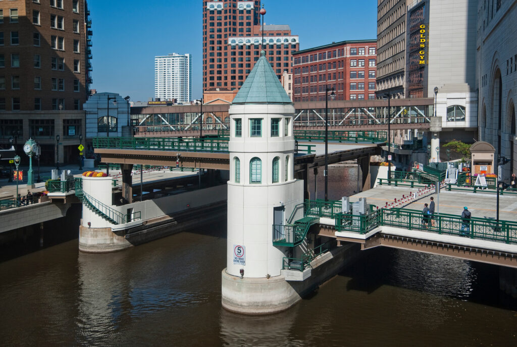 A river with a white turreted control tower, green pedestrian bridges, and surrounding urban buildings in a downtown setting.