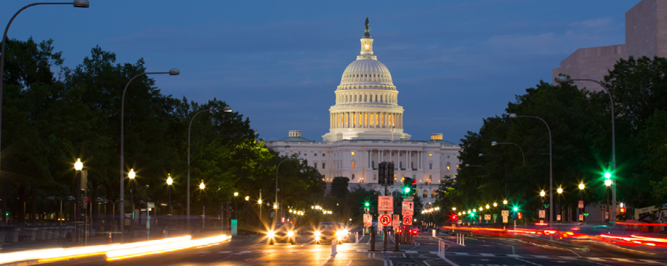 The U.S. Capitol Building is illuminated at dusk, with a view down a busy street showing light trails from moving vehicles.