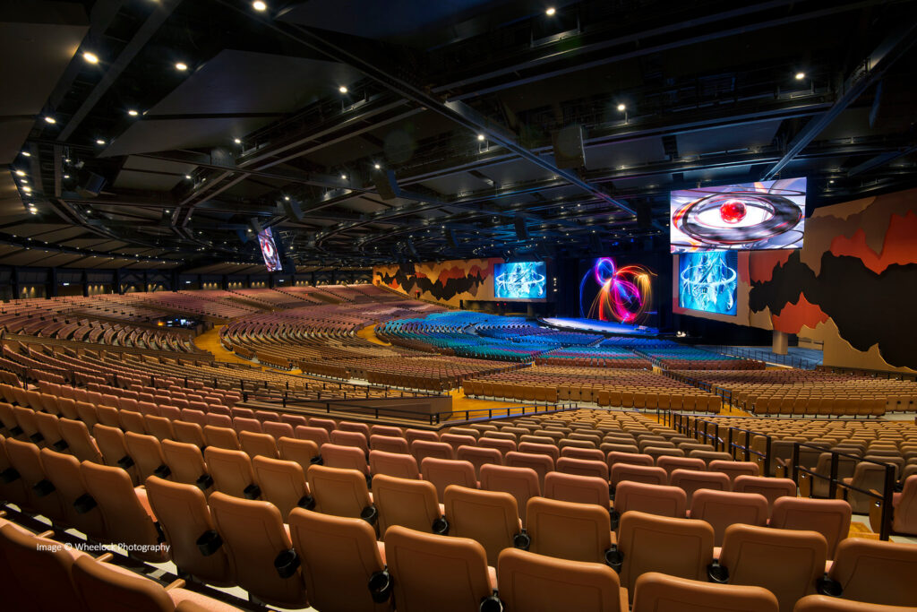 Un grand auditorium vide avec des rangées de sièges beiges, une large scène et plusieurs grands écrans vidéo affichant des visuels abstraits.