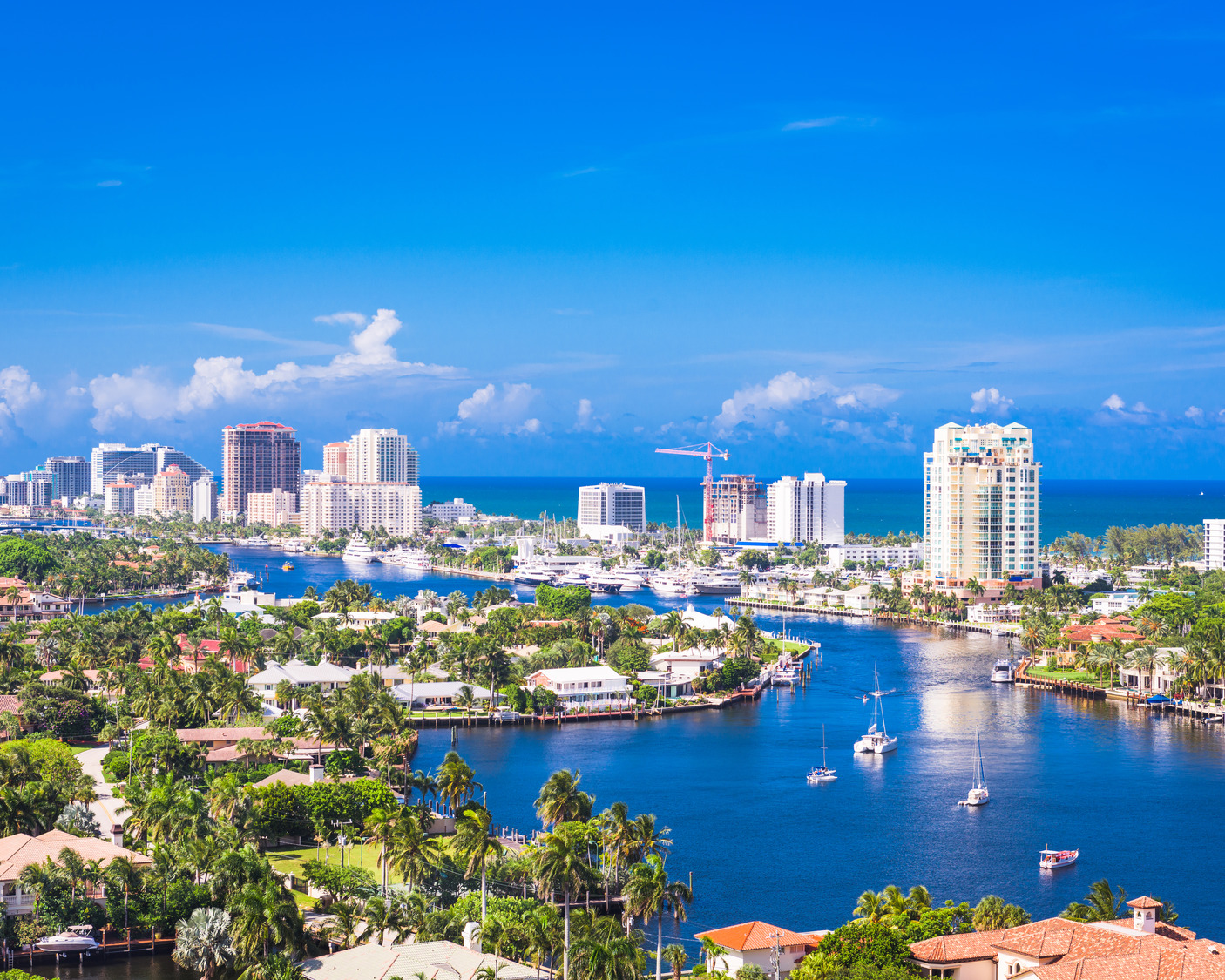 Aerial view of a coastal city with tall buildings, boats in the water, and lush greenery under a clear blue sky.