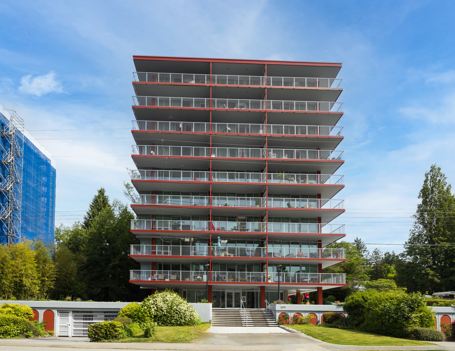 A multi-story modern apartment building with large balconies and red accents stands against a clear blue sky, surrounded by greenery and neighboring buildings.