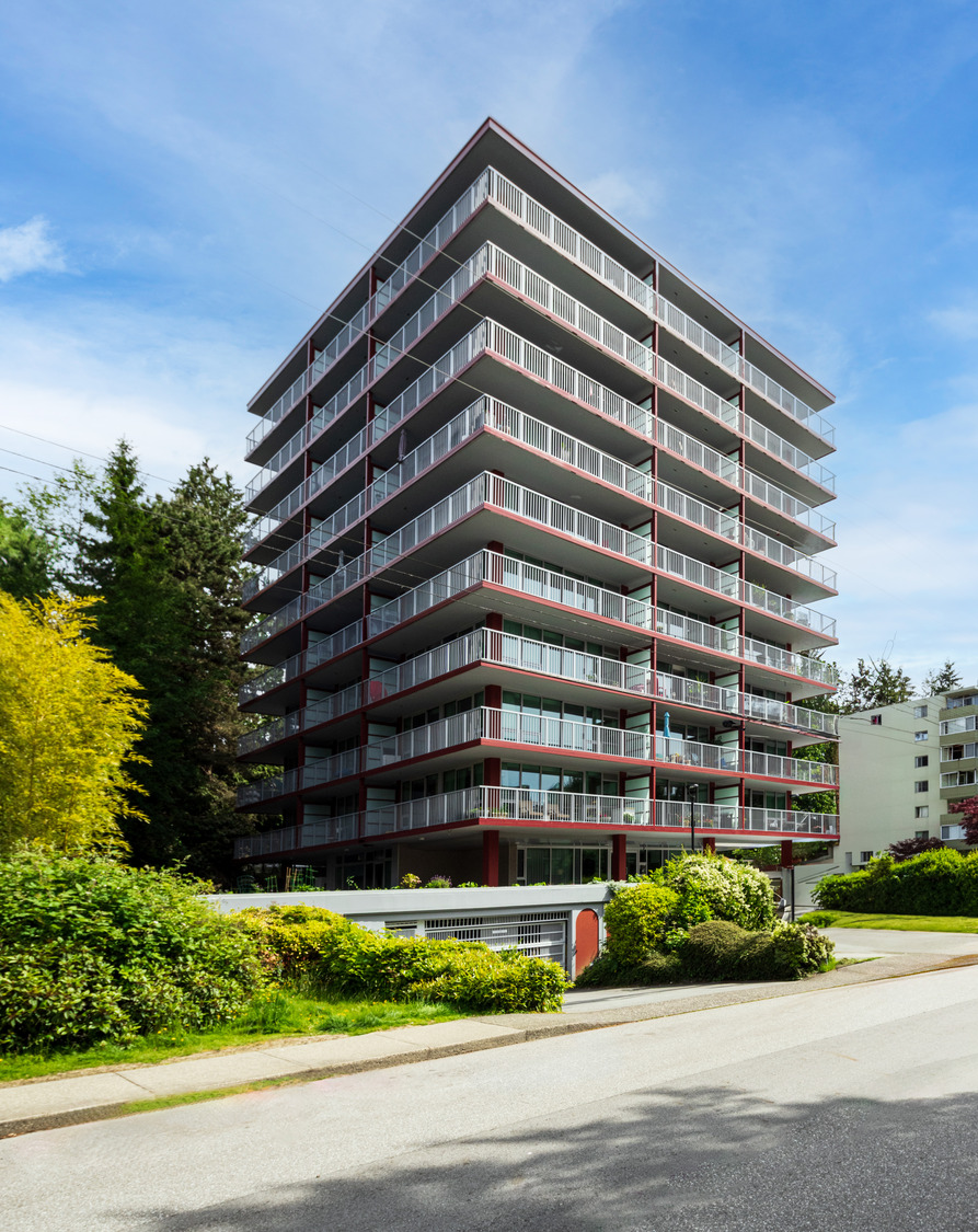 A tall, multi-story residential building with glass balconies on each floor, surrounded by greenery and a few smaller buildings, set against a blue sky.