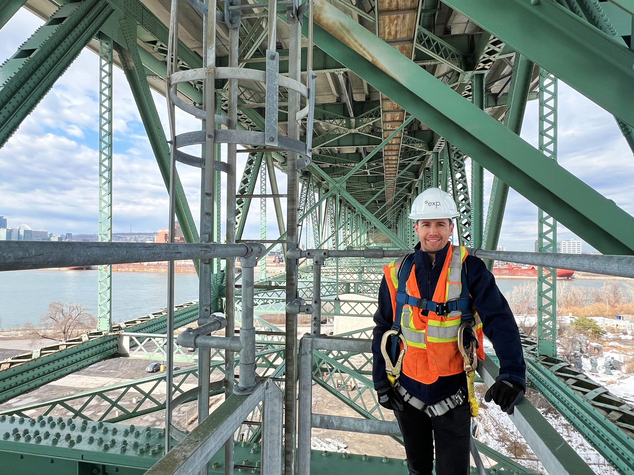 Un ouvrier portant un harnais de sécurité et une tenue de chantier se tient sur une structure d'échafaudage sous un pont métallique vert avec un plan d'eau en arrière-plan.