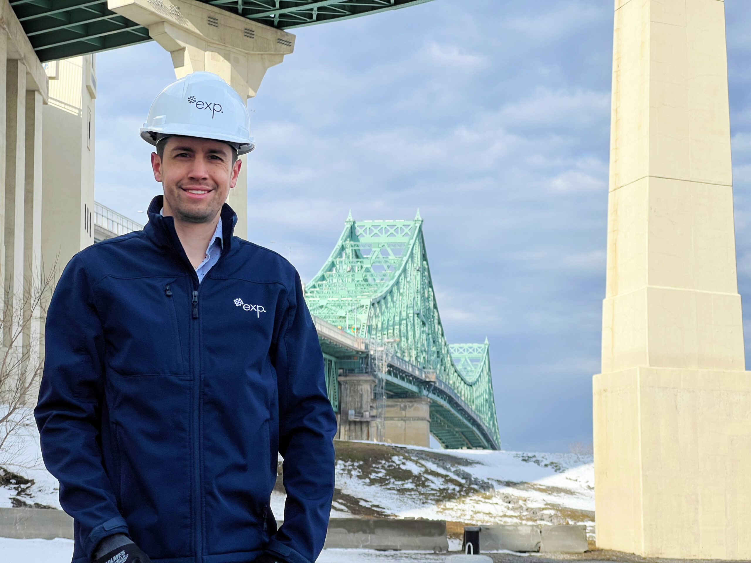 Un homme portant une veste bleue et un casque de chantier blanc se tient dehors près d'un pont par temps nuageux. Le chapeau et la veste portent le logo « exp ». De la neige est visible au sol.