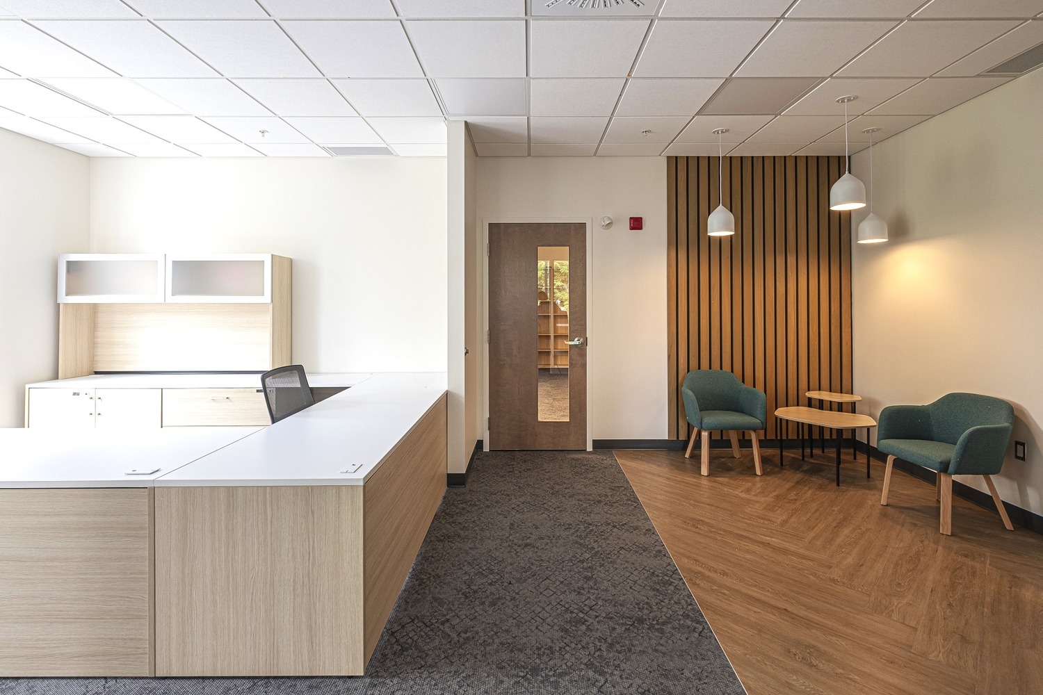 Modern office interior with a wooden reception desk, two green chairs, a small round table, and pendant lights. Walls are cream-colored with a wood panel accent.