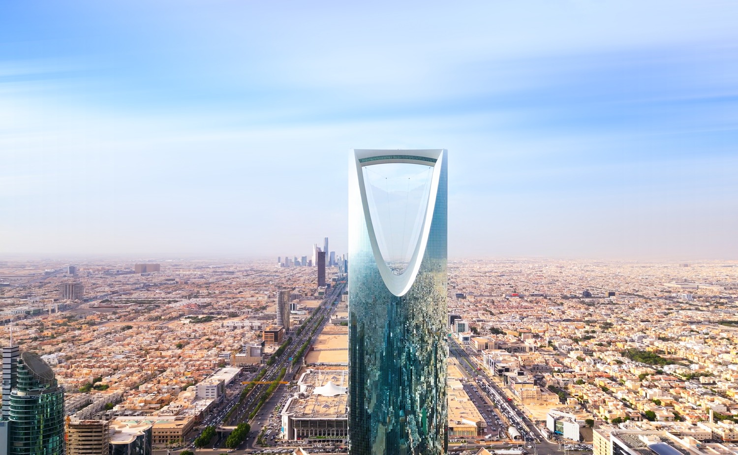 Aerial view of the Kingdom Centre Tower in Riyadh, Saudi Arabia, surrounded by numerous low-rise buildings and wide roads, extending out into the horizon under a clear sky.