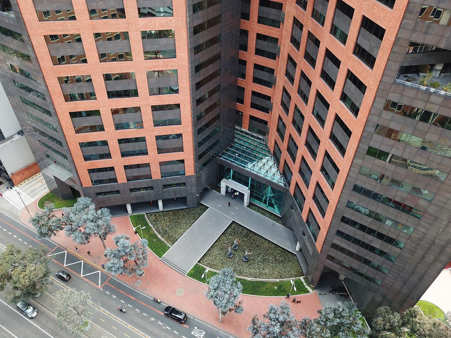 Aerial view of a modern multi-story building with a mix of red and dark exterior finishes, featuring a central courtyard and entrance canopy, surrounded by paved pathways and minimal street traffic.