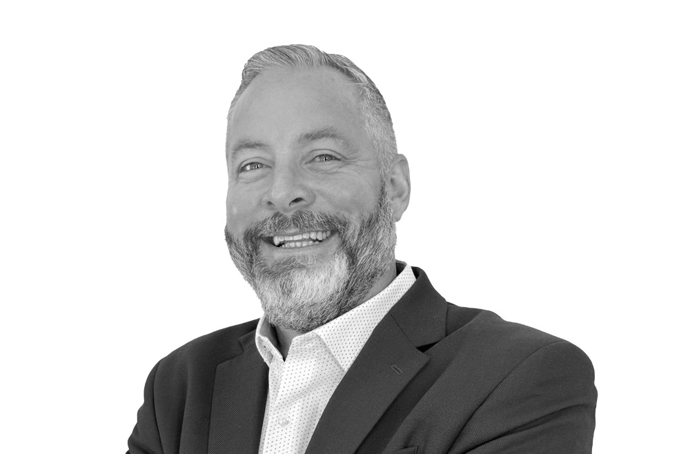 Black and white image of a bearded man with short hair wearing a suit jacket over a patterned shirt, smiling and looking towards the camera.