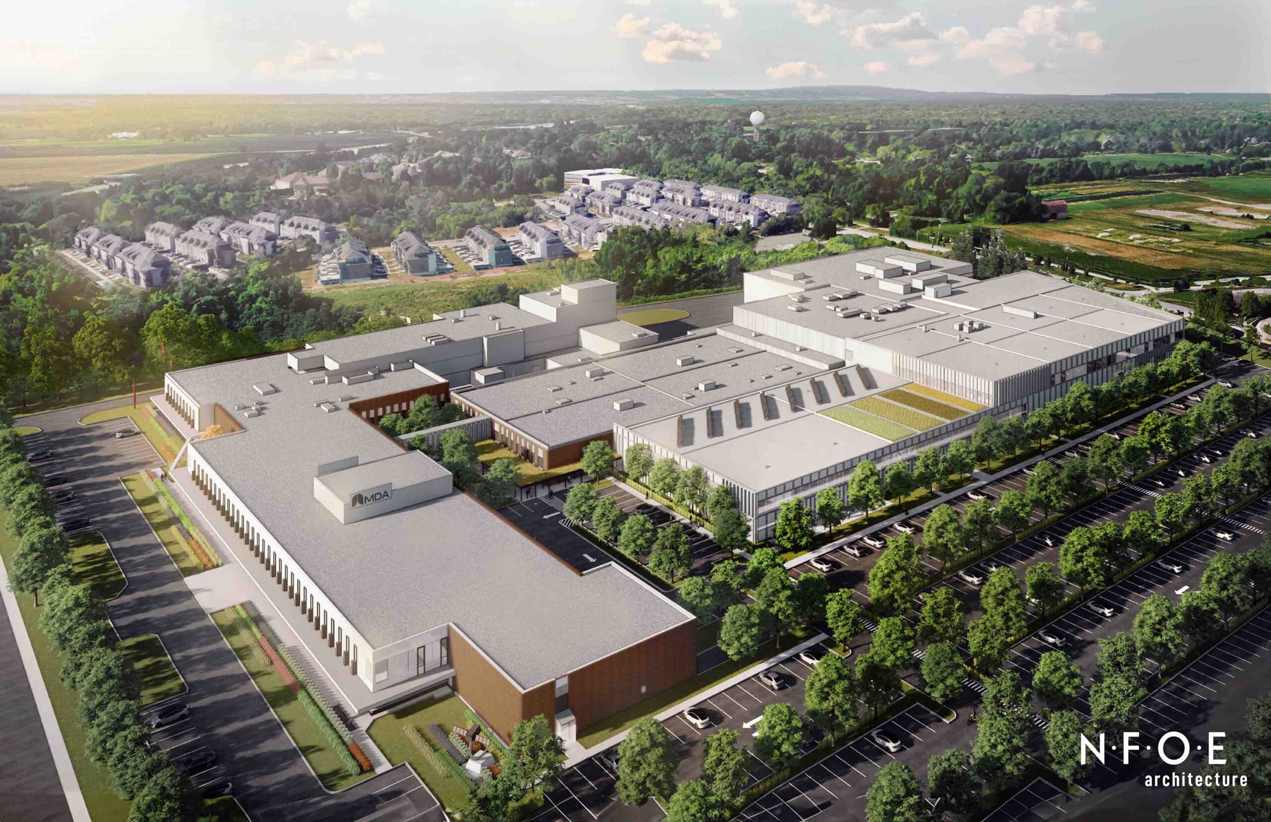 Aerial view of a large industrial complex with multiple buildings surrounded by trees and parking lots, near residential areas and open fields.