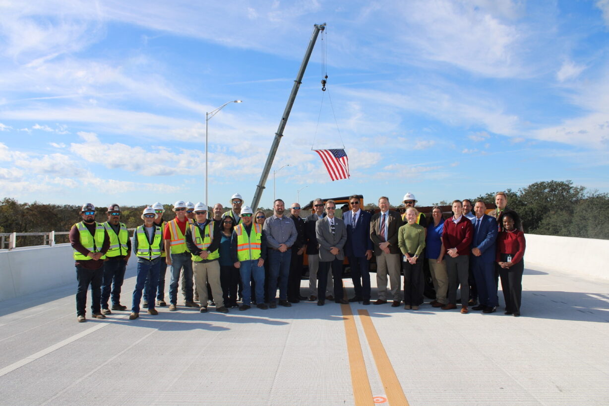 A group of people, including workers in safety vests and individuals in formal attire, stand on a bridge with a crane hoisting the American flag in the background.