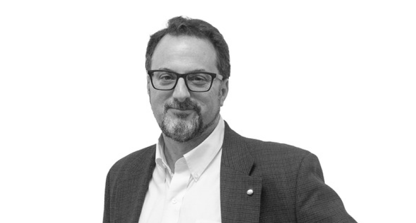 A man wearing glasses and a suit jacket with a white shirt stands against a plain white background.