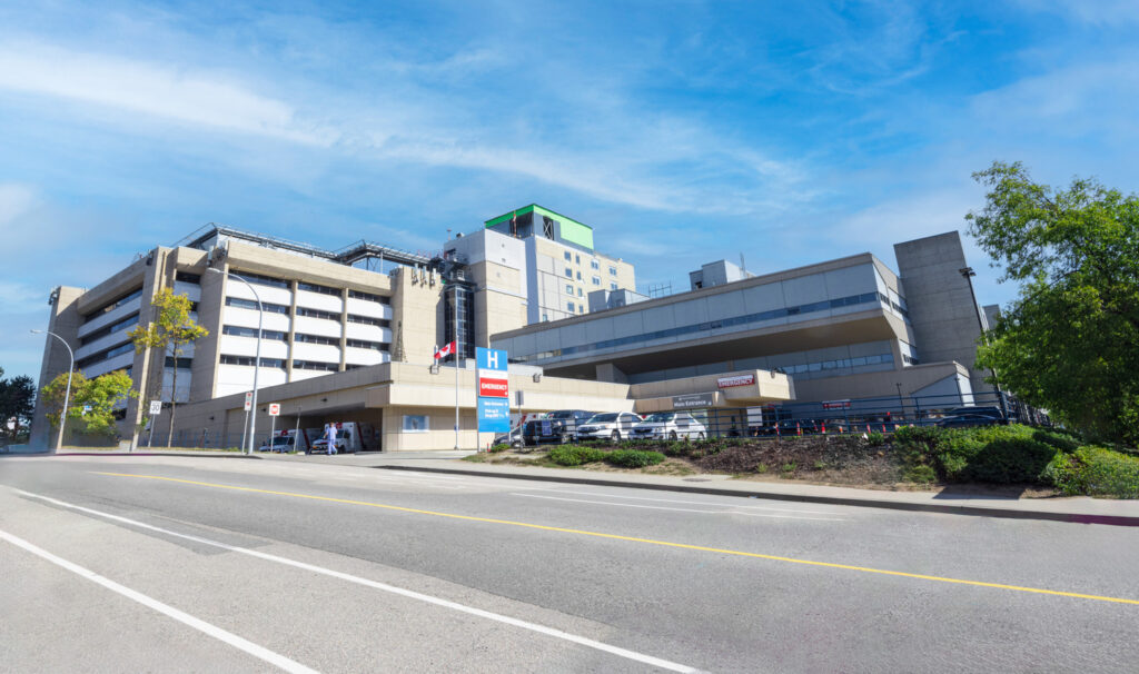 Un grand bâtiment hospitalier avec plusieurs ailes sous un ciel bleu. L'entrée comporte des panneaux d'information sur l'hôpital, avec quelques arbres et une route devant.