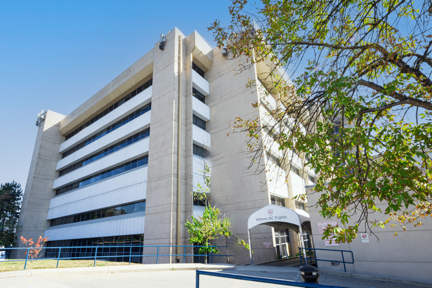 Un immeuble de bureaux moderne en béton avec plusieurs étages et fenêtres. Des arbres et une balustrade sont visibles au premier plan.