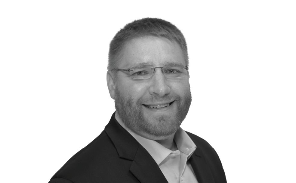 A man with glasses, a beard, and a suit smiles at the camera against a plain white background.