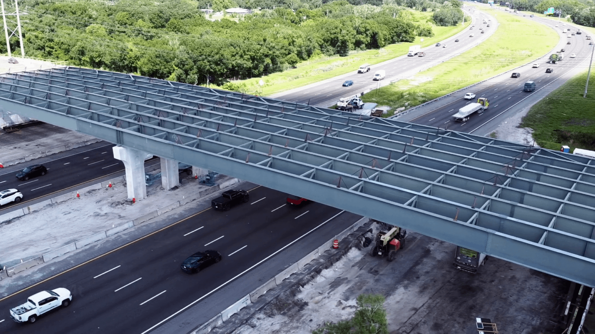 An aerial view of a highway construction site with steel framework for an overpass spanning multiple lanes of traffic.