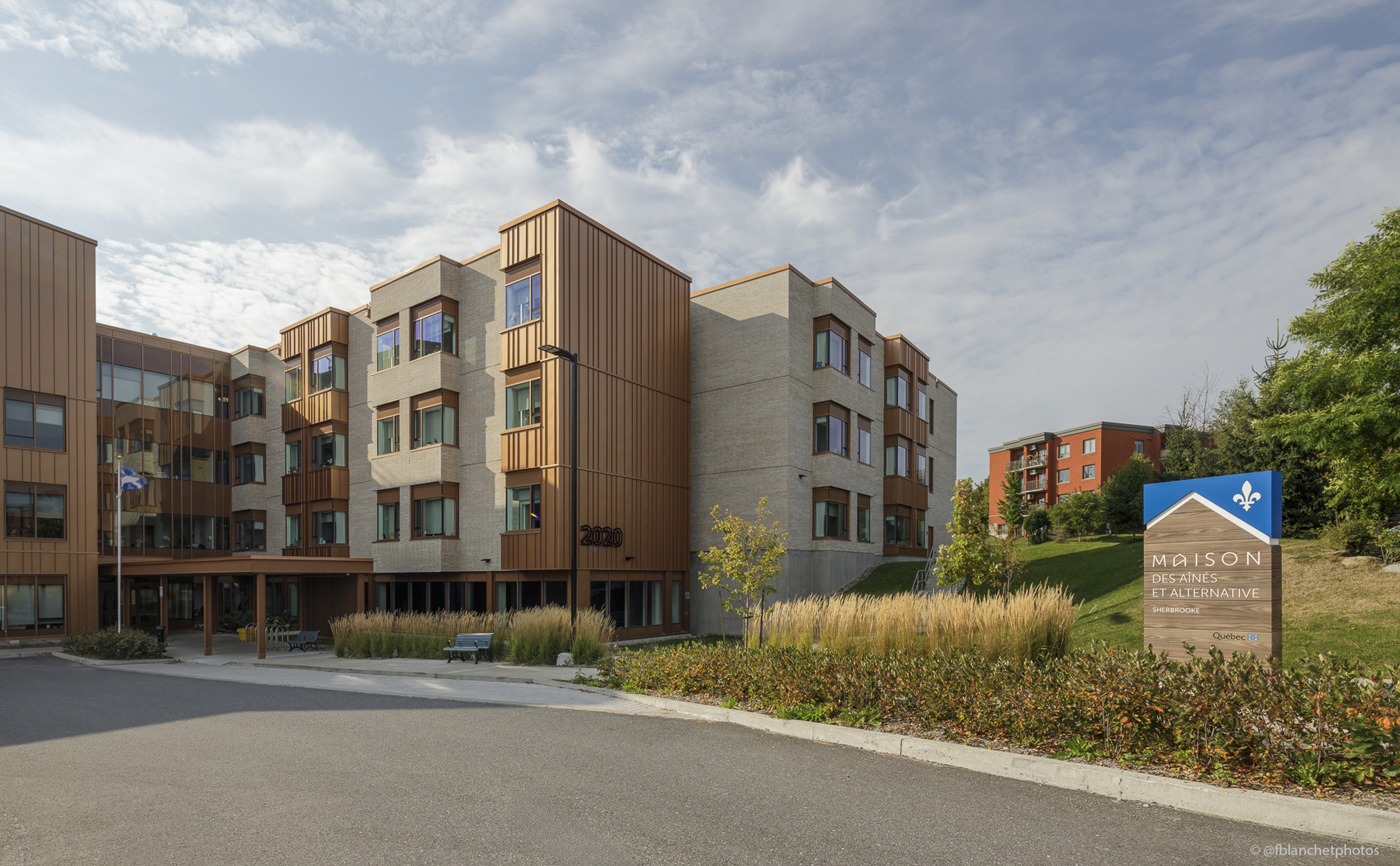 Modern multi-story building with a mix of brick and brown panels, surrounded by landscaping. A sign in front reads "Maison des aînés et alternative.