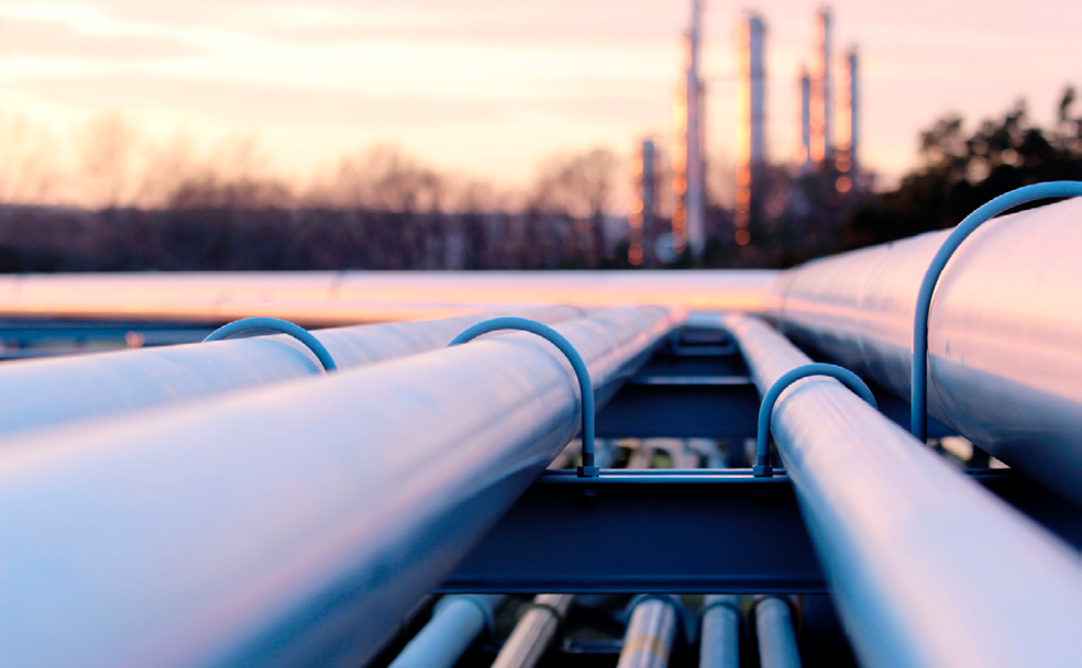 Close-up view of pipeline infrastructure with an industrial facility in the background at sunset.