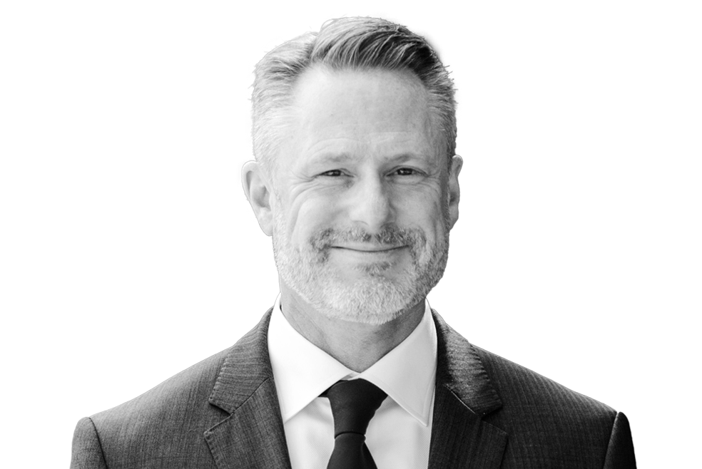 A man with short, gray hair and a beard is smiling, wearing a dark suit and tie in a black and white photograph.