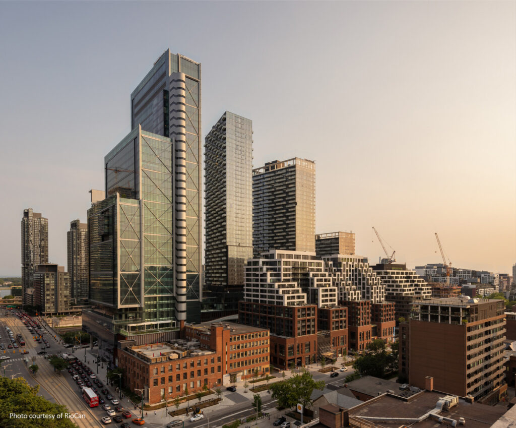 A cityscape featuring modern high-rise buildings, some with geometric designs, alongside older brick structures under a clear sky. Roads with light traffic are visible in the foreground.