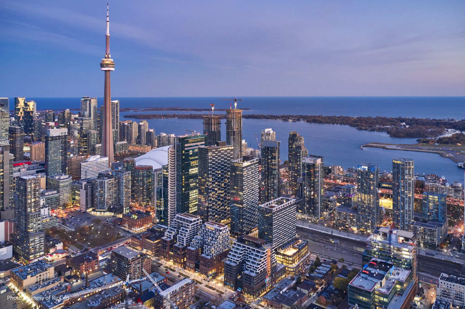 Aerial view of a cityscape with a prominent tower, numerous skyscrapers, and a body of water in the background during twilight.