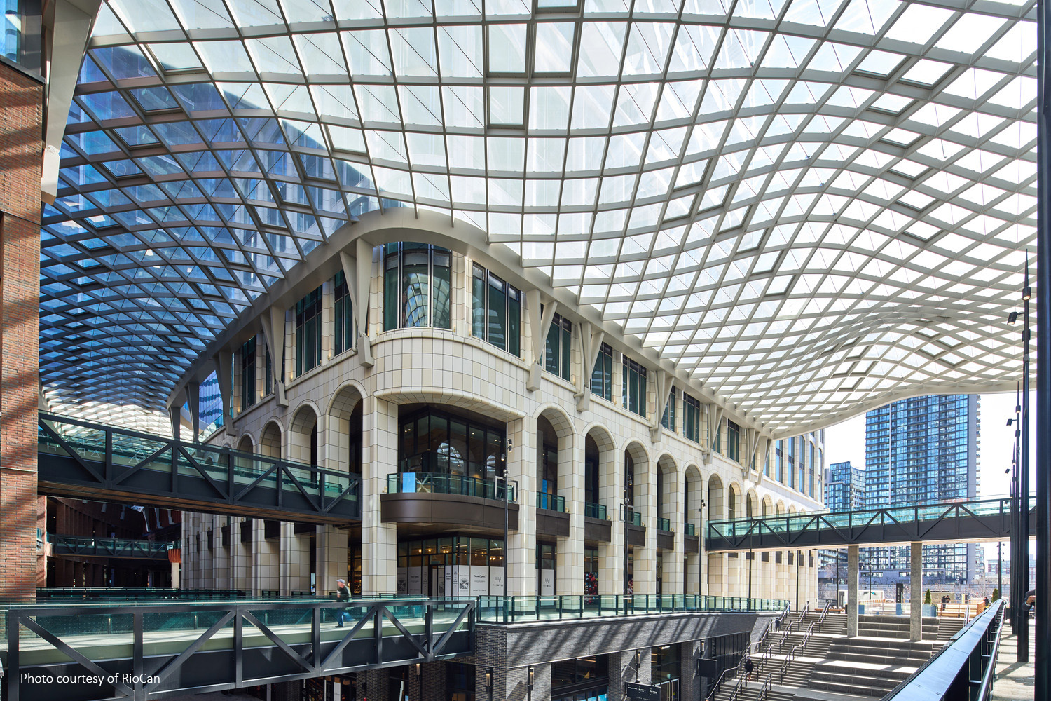 A modern building with a lattice-patterned glass roof and multiple arched windows, featuring pedestrian bridges and a cityscape in the background.