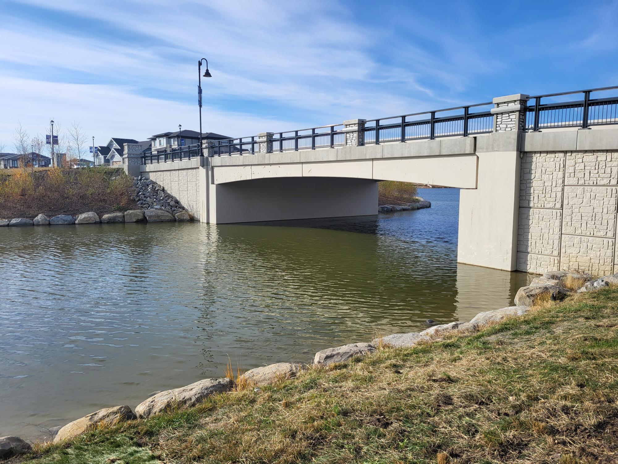 A concrete bridge spans over a calm body of water, with a lamp post on top. Rocks and grass are in the foreground, and houses are in the background under a blue sky.