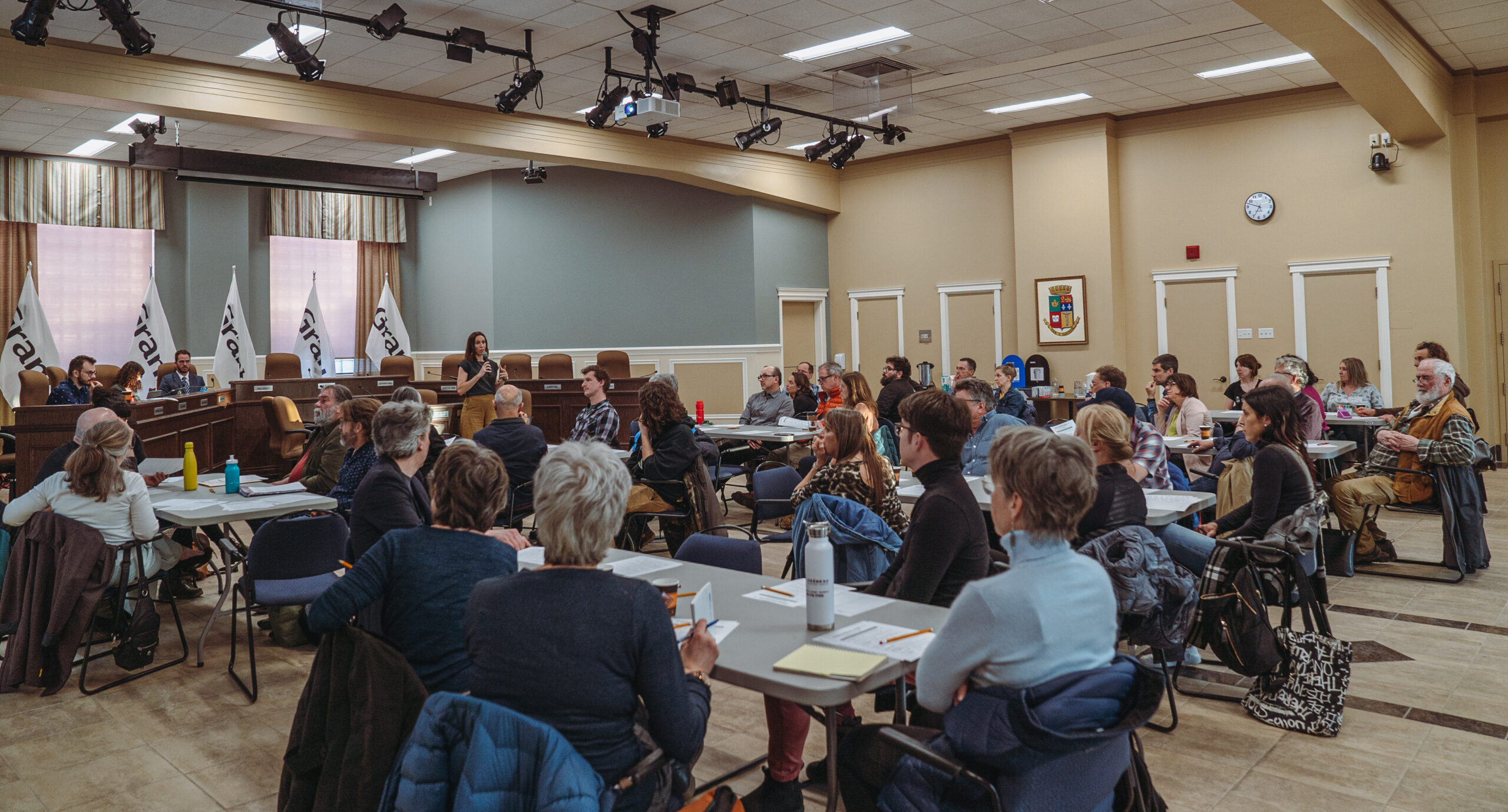 Un groupe de personnes assises dans une salle de conférence avec un orateur à un podium et un panel de fonctionnaires en arrière-plan.