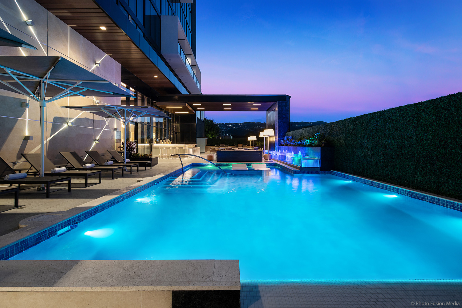 Un espace piscine extérieur moderne au crépuscule, avec des chaises longues, des parasols et une piscine éclairée avec un jeu d'eau. Un bâtiment et une haie forment la toile de fond, avec une vue sur les collines à l'horizon.