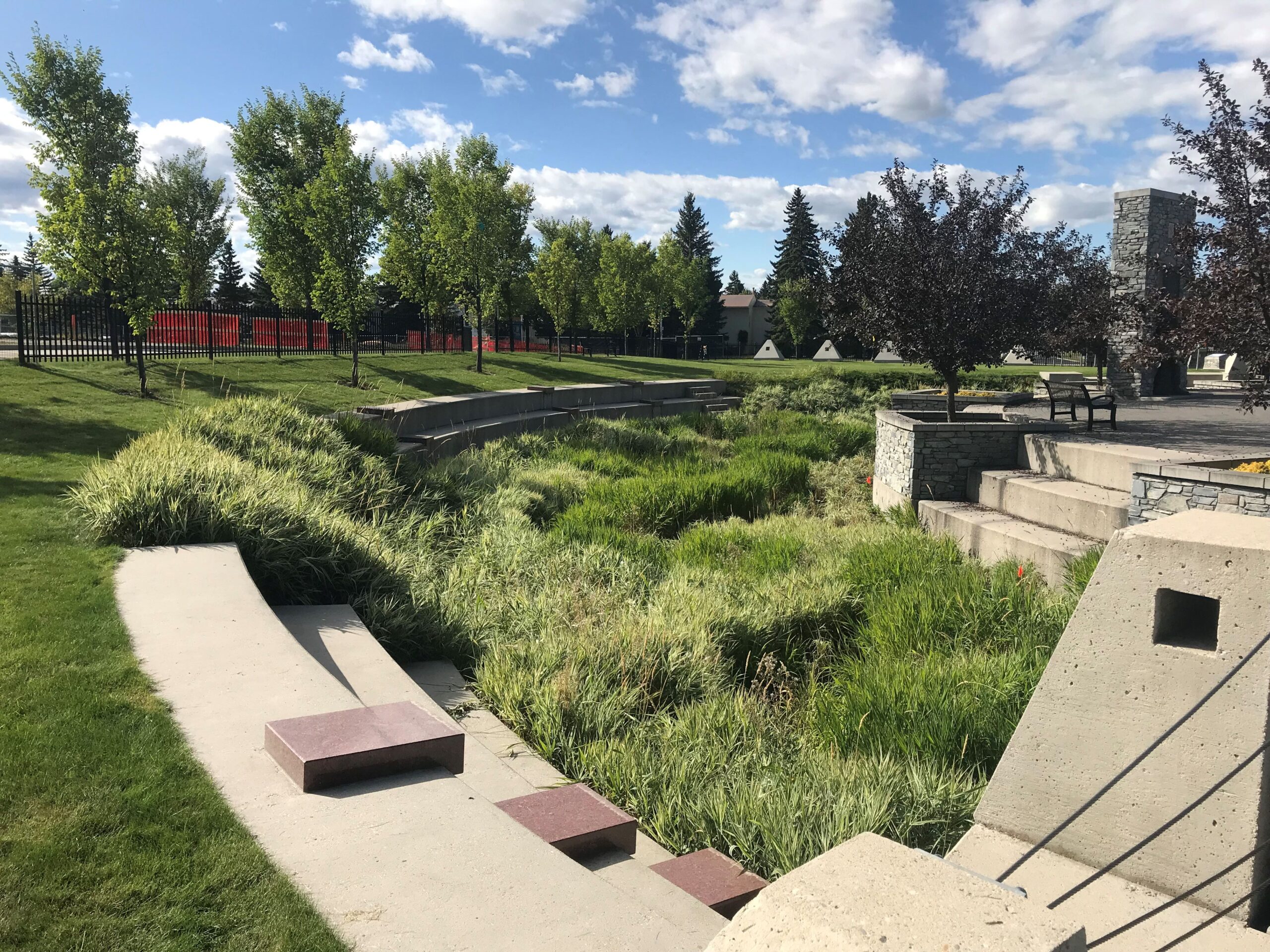 Outdoor park area with paved walkways, steps, and green grassy sections. Trees line the area under a partly cloudy sky.