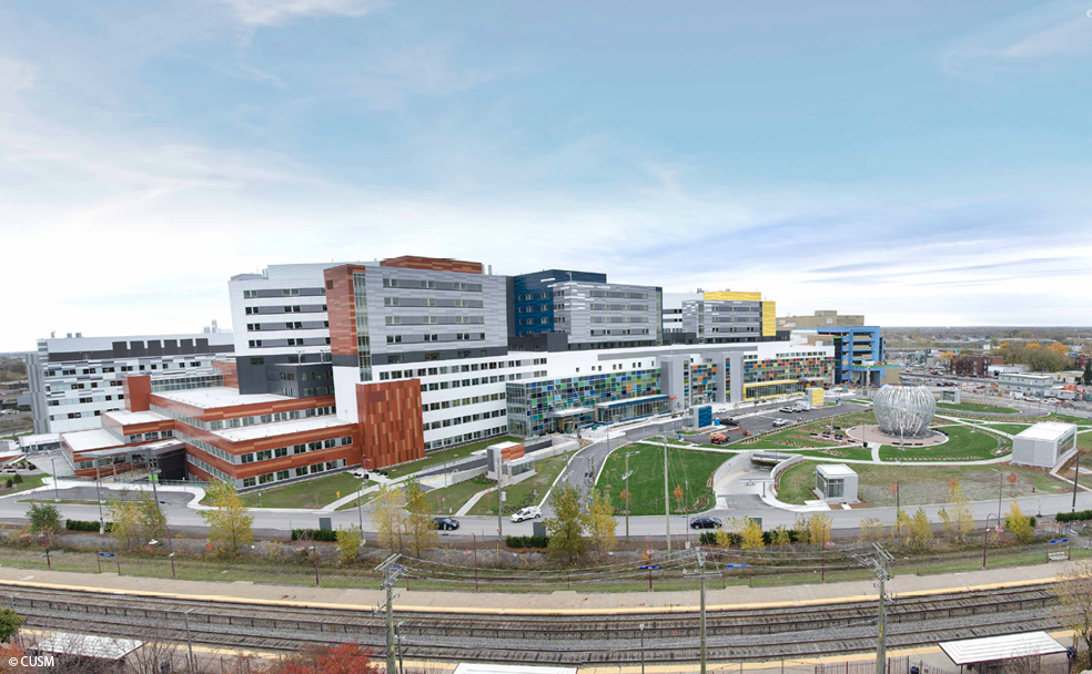 Un grand complexe hospitalier à l'architecture moderne, entouré de pelouses et d'arbres, sous un ciel partiellement nuageux. Une ligne de chemin de fer est visible au premier plan.