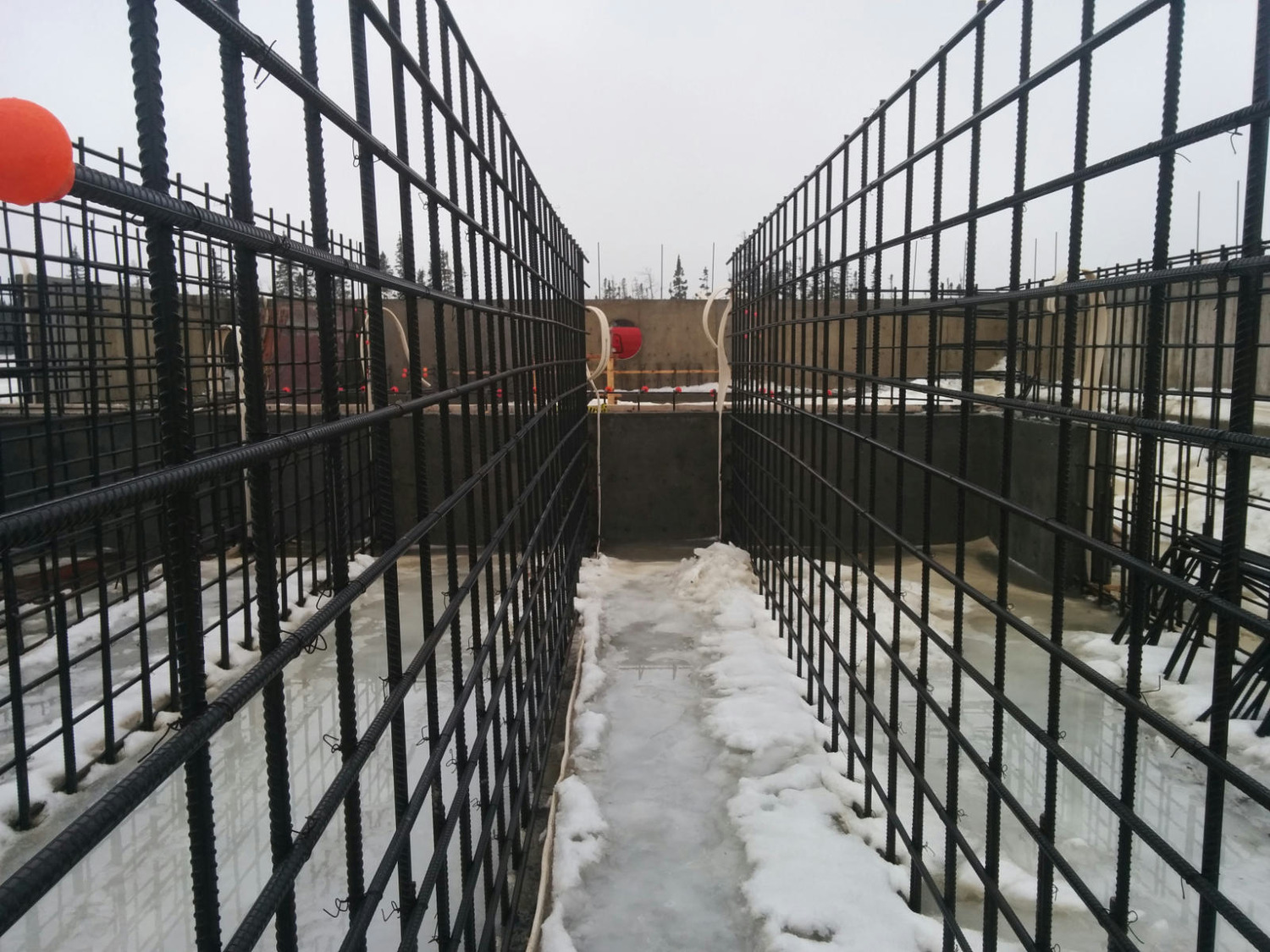 Metal rebar framework for concrete construction with snow on the ground and orange safety caps on protruding bars. Overcast sky in the background.