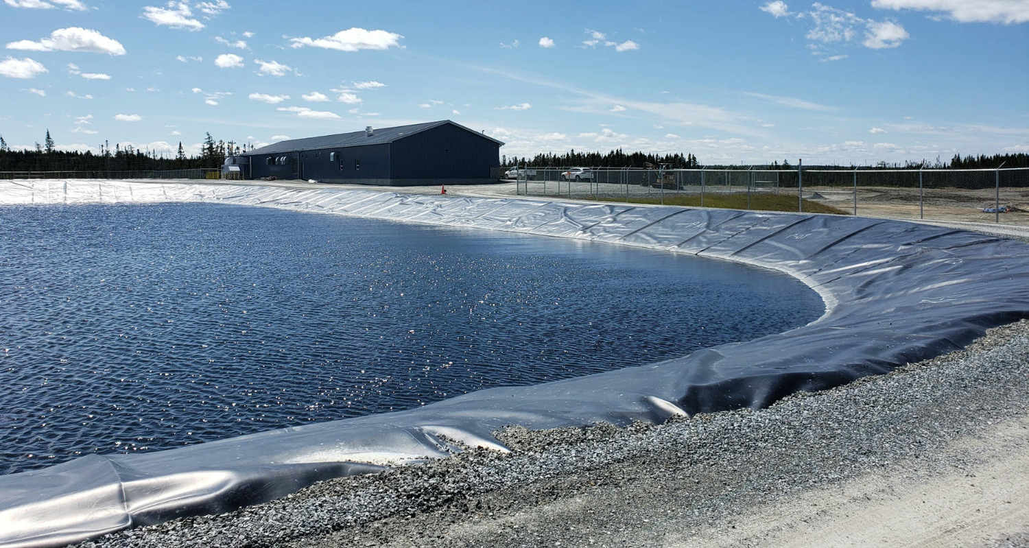 A large, lined water retention pond near a fenced industrial building under a blue sky with scattered clouds.