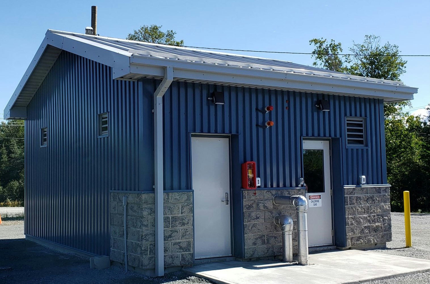 A small, blue corrugated metal utility building with a sloped roof. It has two doors, a red fire extinguisher, and multiple vents. Trees and a clear sky are in the background.