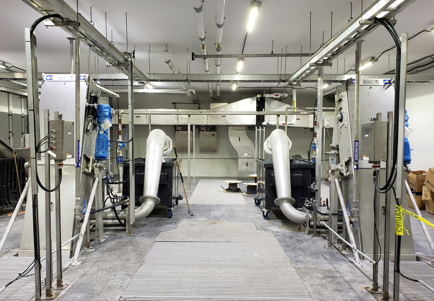 Industrial facility with large metal machinery, pipes, and overhead lighting in a spacious room. Safety tape is visible on the right side.