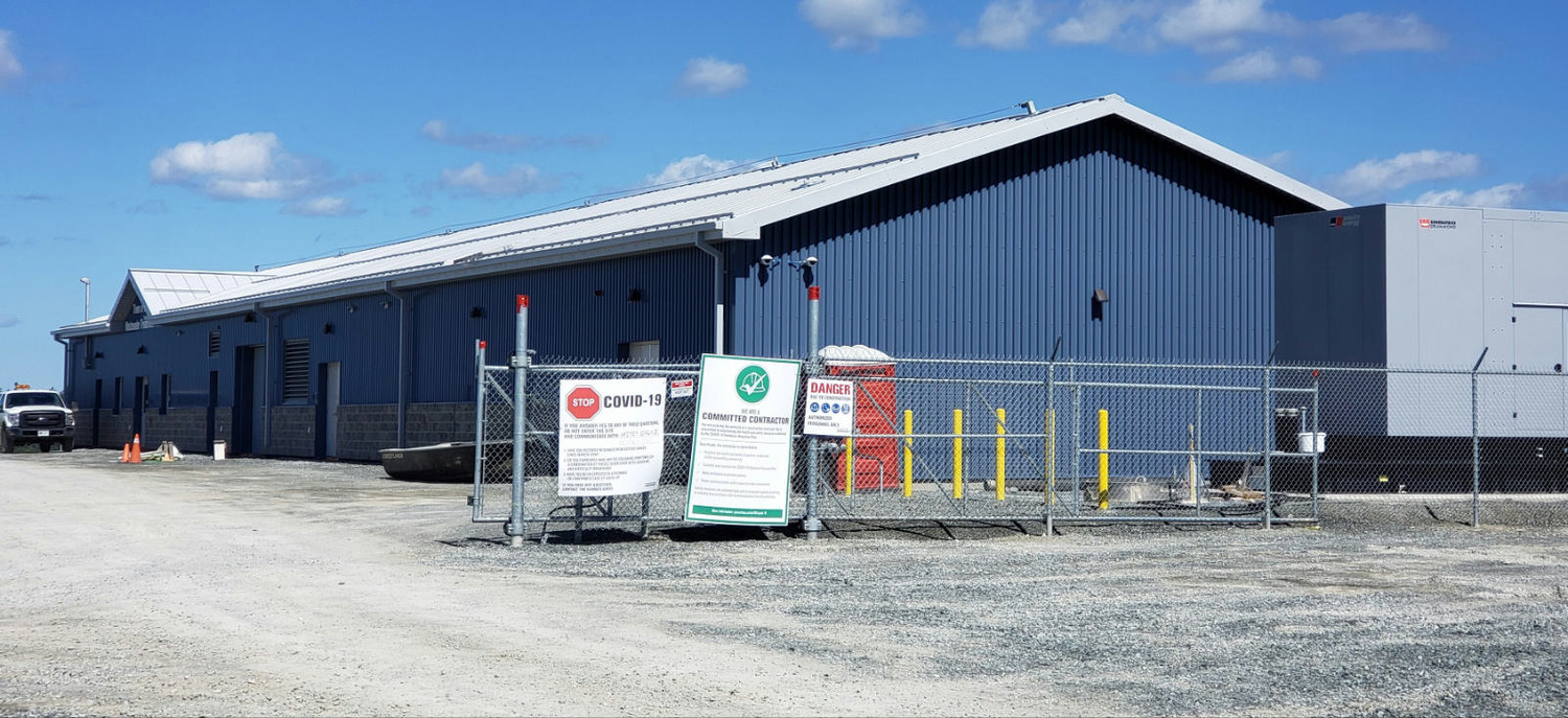 A large blue industrial building with a white roof is surrounded by a chain-link fence. Signs on the fence provide safety and COVID-19 information. A clear sky is in the background.