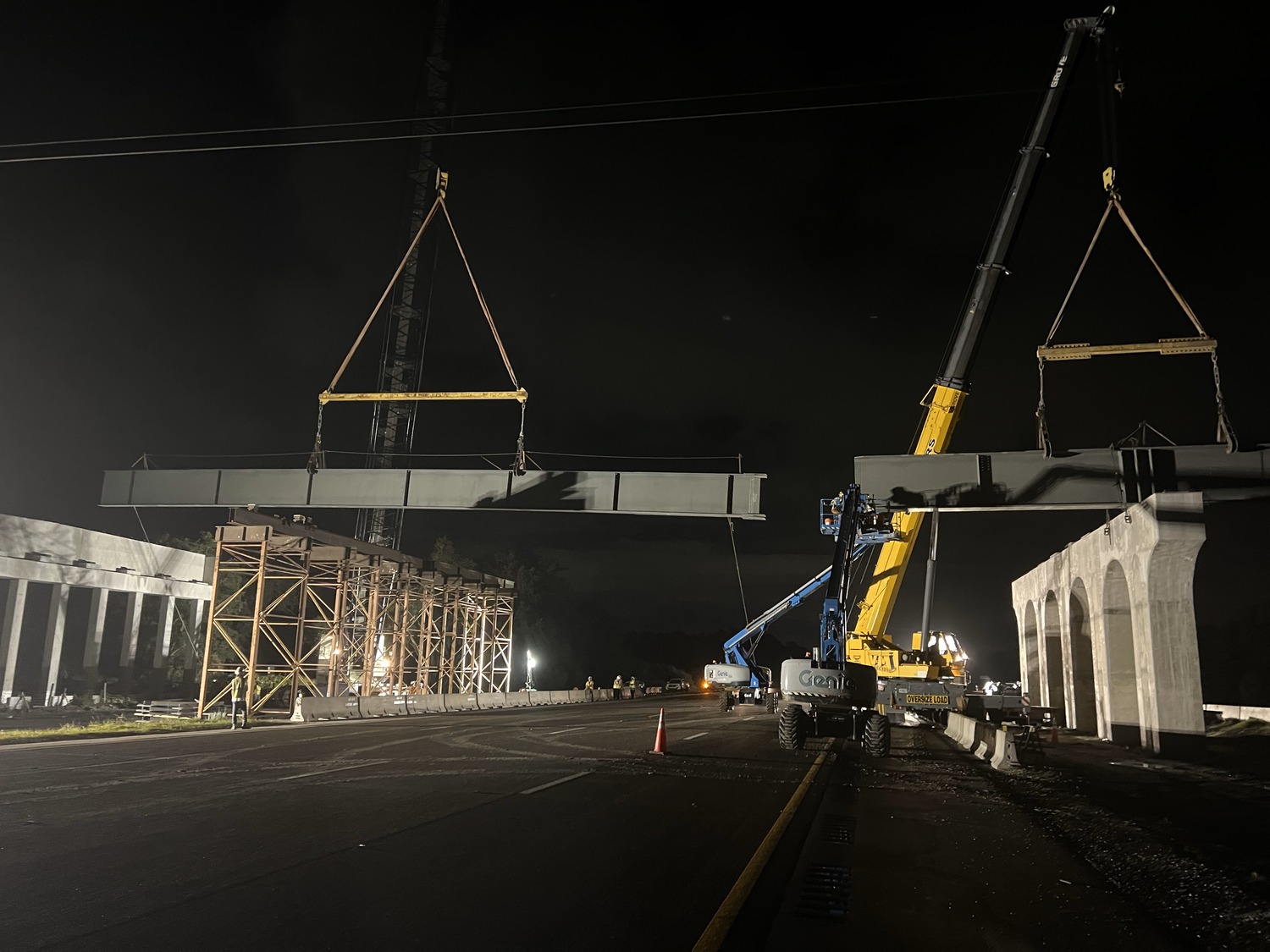 Construction site at night with cranes lifting large beams to assemble a bridge. Workers and equipment are visible under lights.