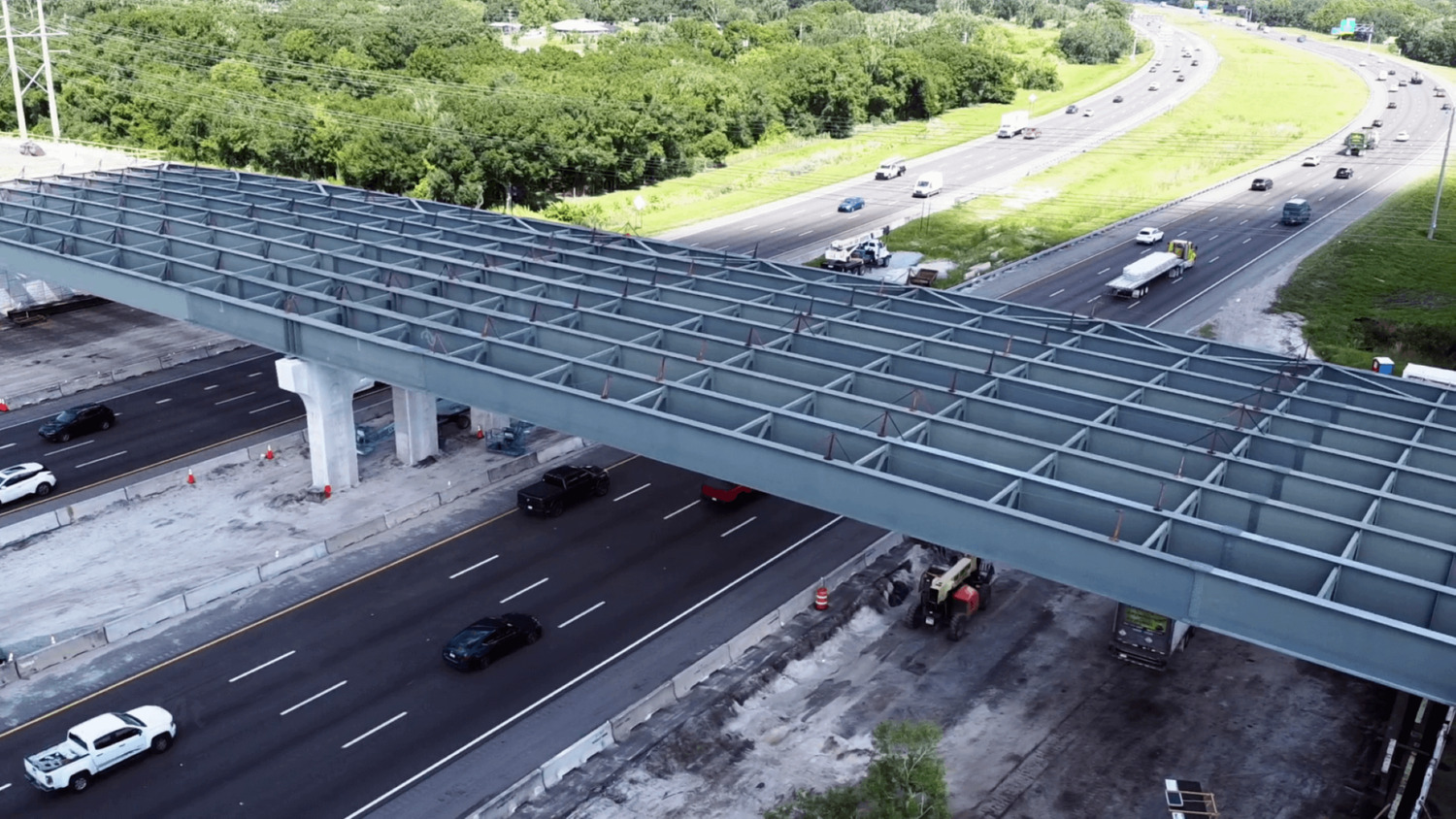 Aerial view of an elevated highway under construction over a busy road. Traffic flows below with vehicles visible. Workers and construction equipment are present on site.