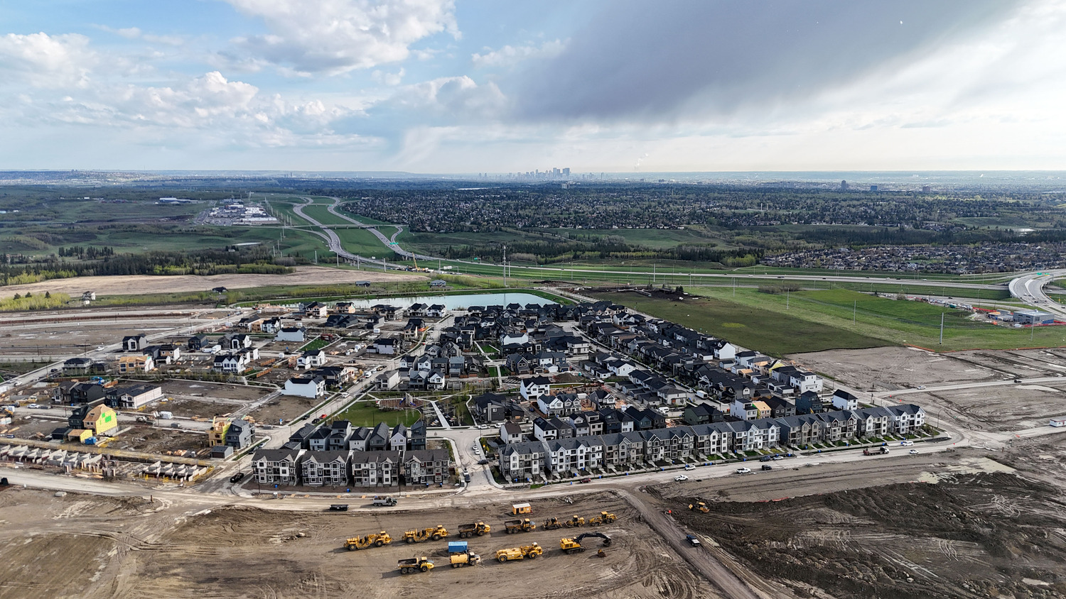 Aerial view of a housing development surrounded by open land, roads, and construction vehicles, with a distant city skyline under a partly cloudy sky.