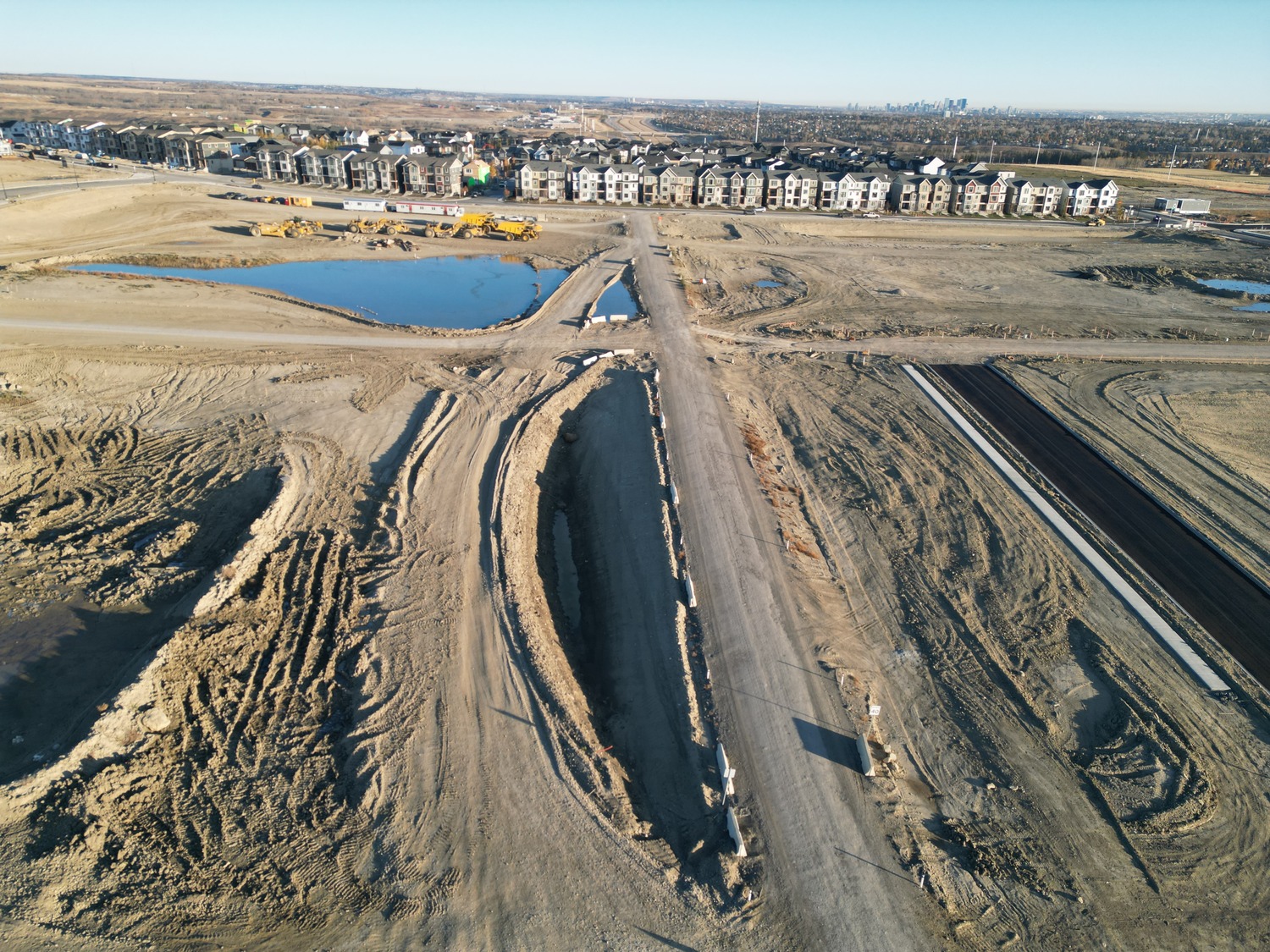 Aerial view of a construction site with dirt roads, equipment, and partially completed infrastructure. Residential buildings are visible in the background.